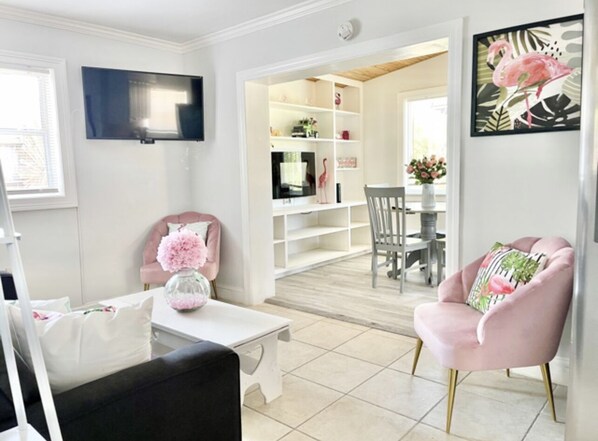 A modern living room with a TV, a pink armchair, a coffee table with a vase, and a dining area in the background.