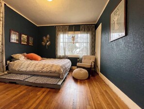 Ein Schlafzimmer mit einem Bett, einem Sessel, einem Fenster mit Vorhängen und einer Wand mit gerahmten Bildern.