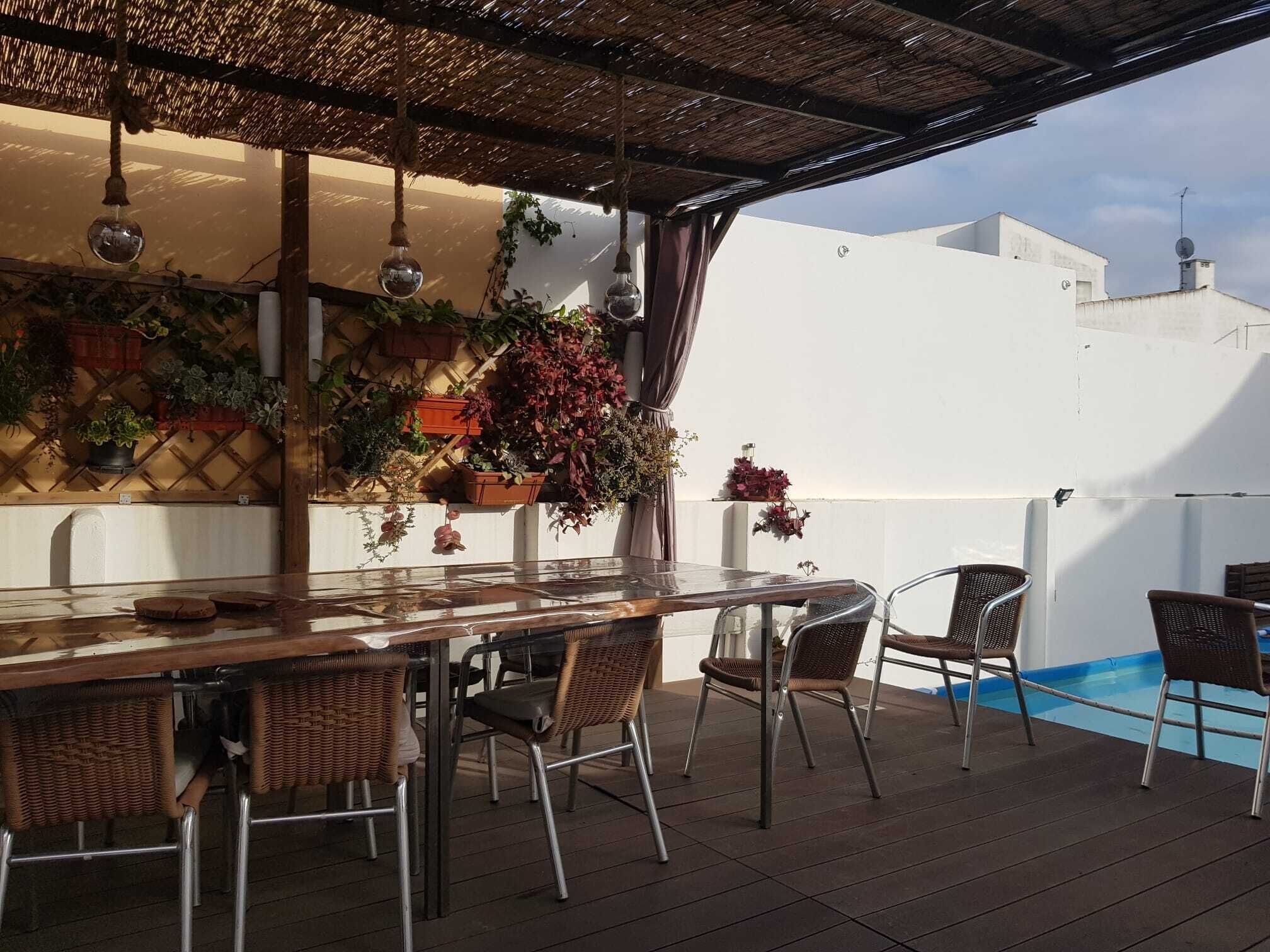 An outdoor patio with a wooden table and wicker chairs, surrounded by potted plants and hanging light fixtures.