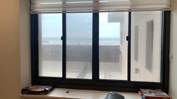 A room with a large window overlooking a beach. There are board games on a shelf below the window. - Beach Getaway (Long Beach)
