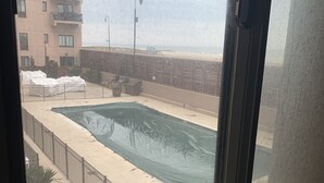 A view of a swimming pool with a metal fence, a beach, and a building in the background. - Beach Getaway (Long Beach)