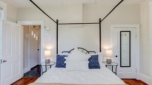 Ein ordentlich eingerichtetes Schlafzimmer mit einem Himmelbett, zwei Nachttischen mit Lampen, einer Tür mit Spiegel und einem Flur, der in einen weiteren Raum führt.