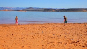 Ein Strand mit zwei Personen, einem Kind und einem Erwachsenen, in der Nähe ruhigen Wassers mit Bergen im Hintergrund.
