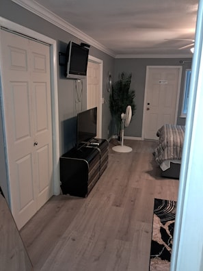 A room with a flat-screen TV mounted on the wall, a black entertainment unit, a standing fan, and a door with a sign.