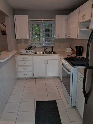 A compact kitchen with white cabinets, a stove, a sink, and a window with blinds. - The Jones Suites (Freeport)