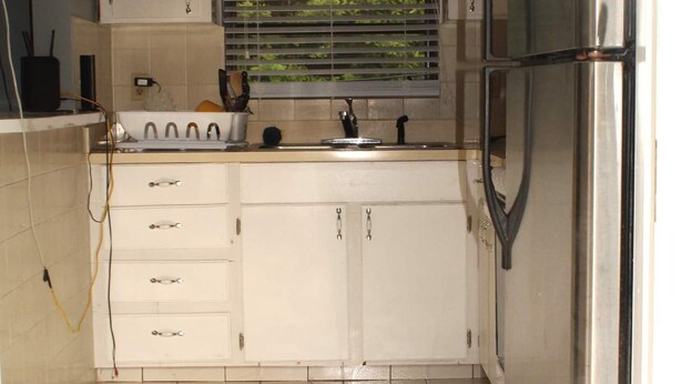 A compact kitchen with white cabinets, a stainless steel refrigerator, and a window with blinds. - The Jones Suites (Freeport)