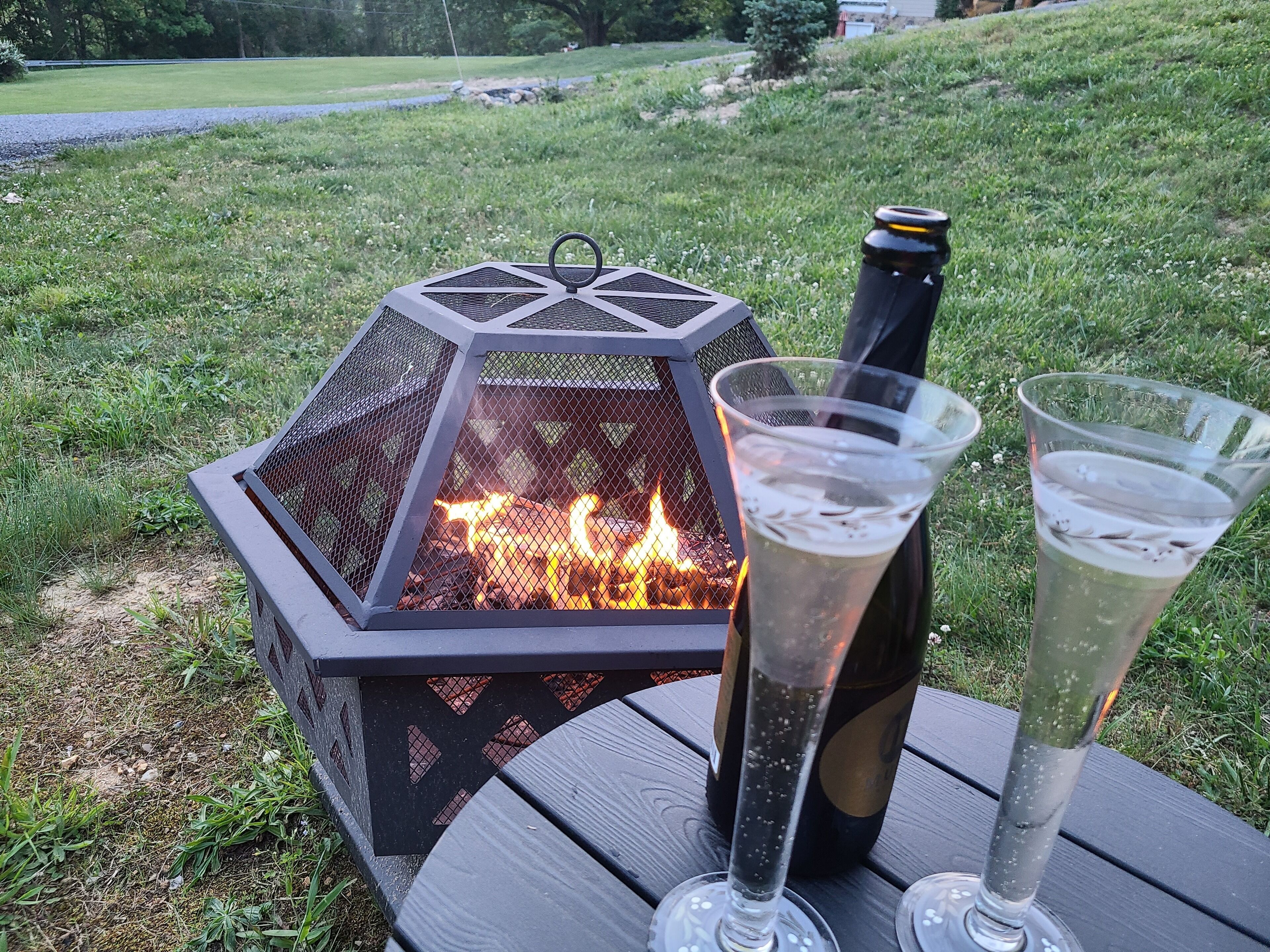 A hexagonal fire pit with a bottle of champagne and two glasses on a wooden table.