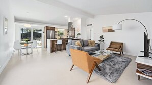 Ein modernes Wohnzimmer mit Sofa, Couchtisch und Sesseln. Im Hintergrund ist eine Küche mit Essbereich zu sehen.