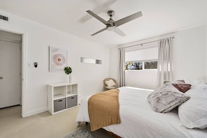 A bedroom with a bed, a ceiling fan, a window with curtains, a bedside table, and a wall-mounted shelf.