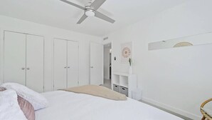 Ein Schlafzimmer mit einem Bett, einem Deckenventilator, weißen Kleiderschränken, einem kleinen weißen Regal mit einer Vase und einem Korbstuhl.