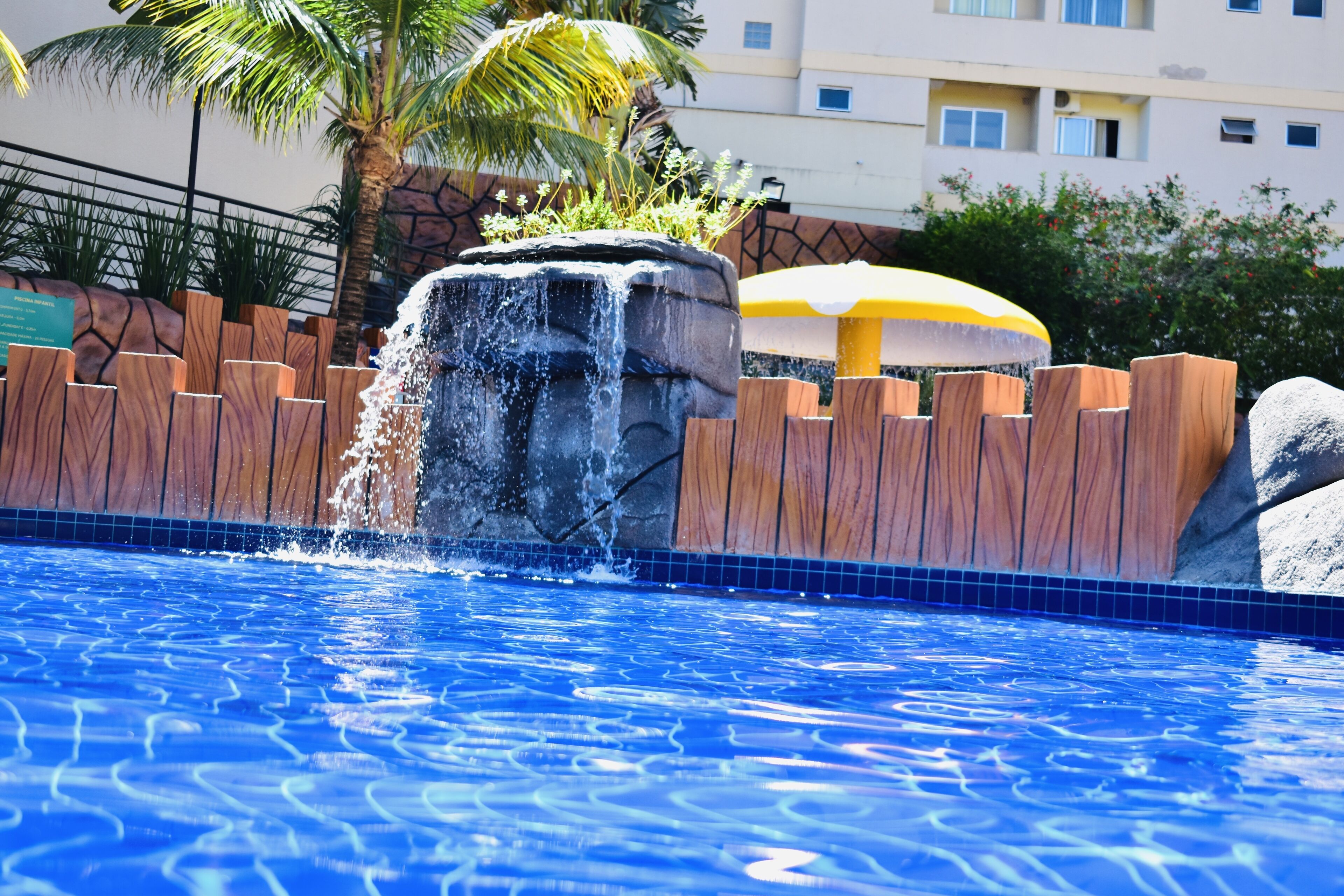 A swimming pool with a waterfall feature, surrounded by wooden fencing and palm trees.