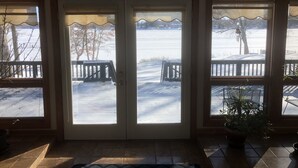 En vy genom ett par glasdörrar som leder ut till ett snötäckt utomhusområde med en bänk och ett bord.