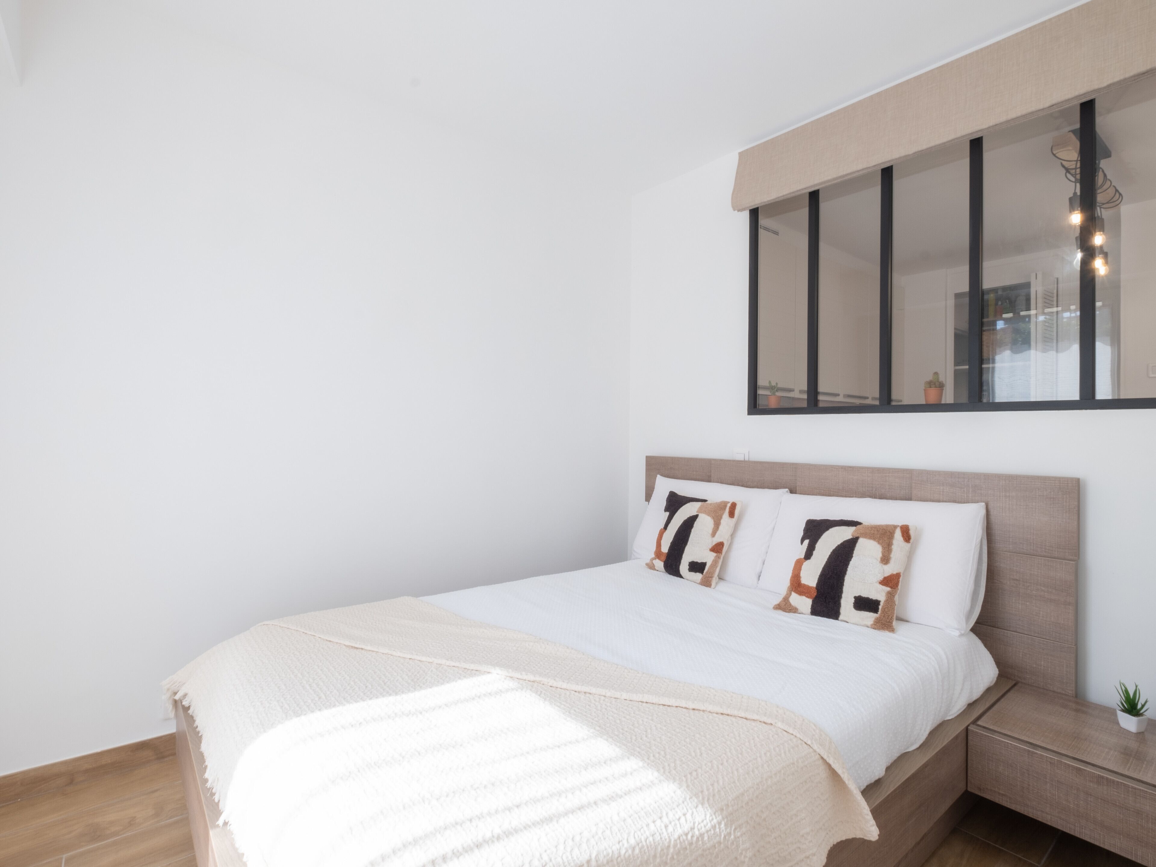 A modern bedroom with a large bed, a nightstand, a small plant, and a window with blinds.