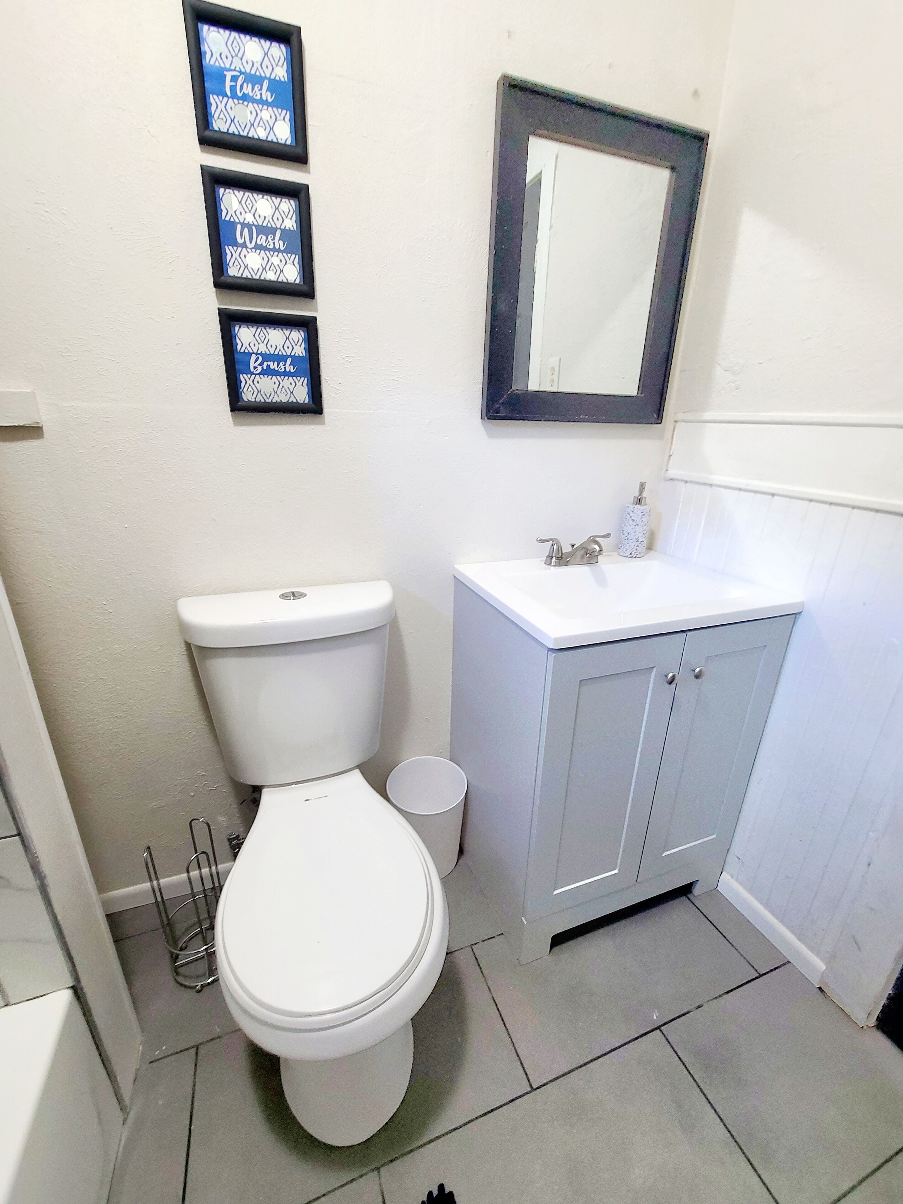A bathroom with a toilet, sink, and mirror. There are three framed signs above the toilet.