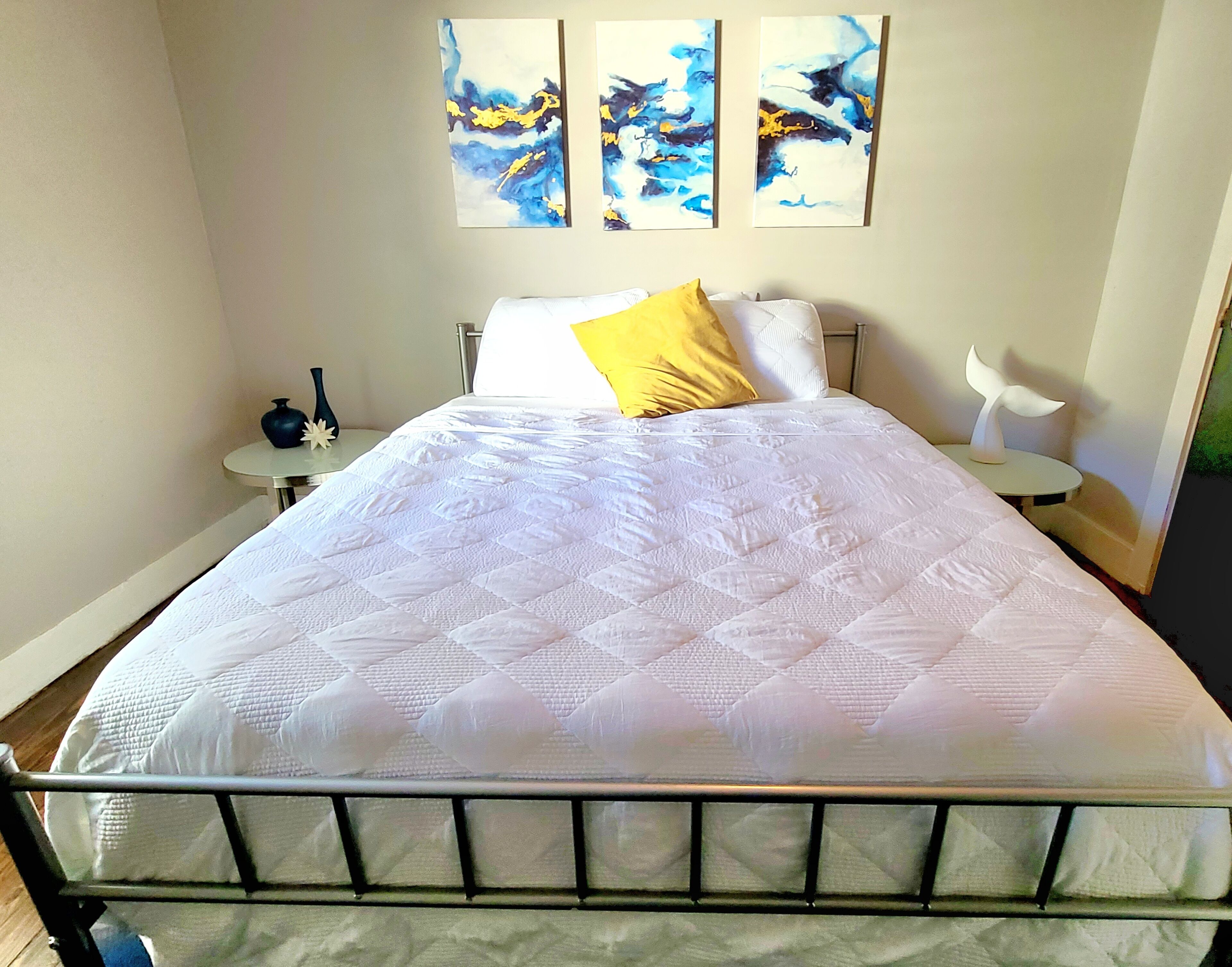 Een netjes opgemaakt bed met een witte quilt, een geel kussen en een metalen hoofdbord. Daarboven hangen drie abstracte schilderijen aan de muur.