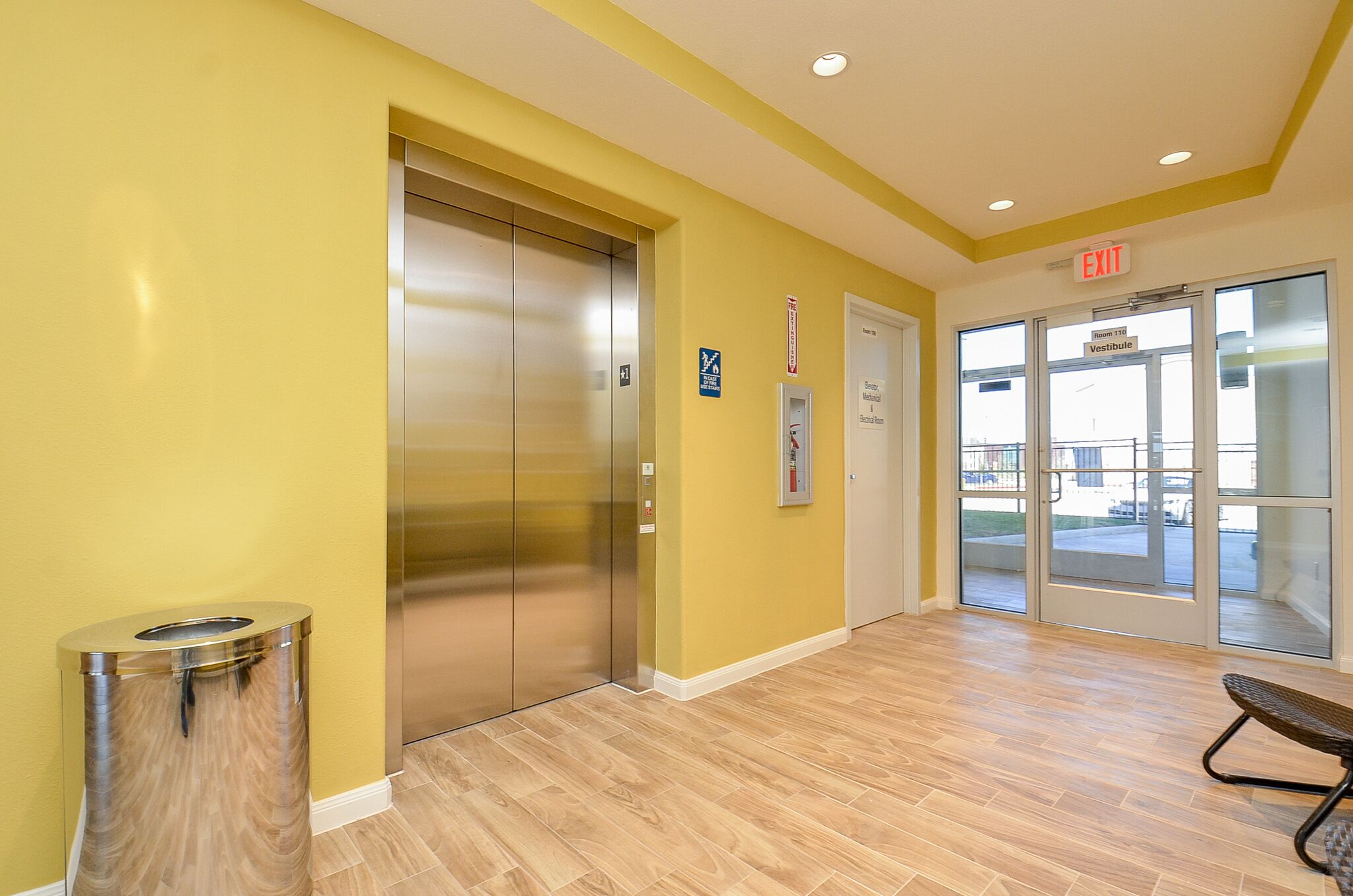 An elevator, a trash can, and a bench in a hallway with yellow walls and wooden flooring.