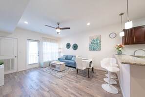 Un apartamento moderno con comedor, sala con sofá azul, ventilador de techo y cocina con gabinetes de madera.