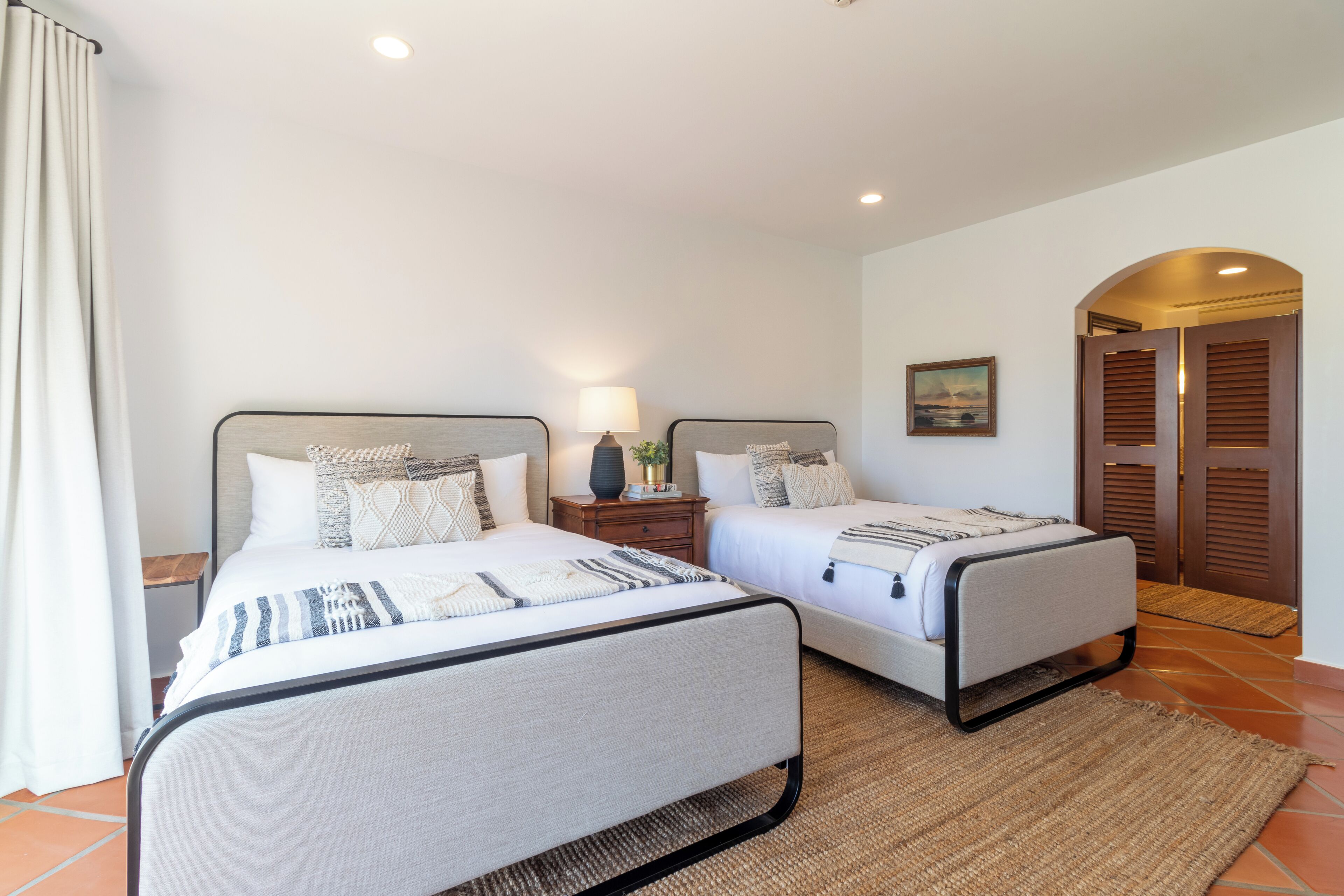 A double room with two beds, a nightstand with a lamp, a painting on the wall, and a wooden door.