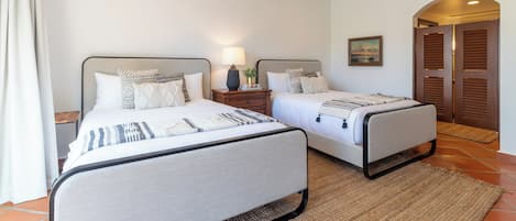 Quarto duplo com duas camas, uma mesa de cabeceira com abajur, um quadro na parede e uma porta de madeira.