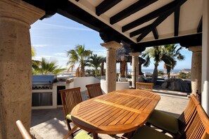 Een terras buiten met een houten tafel en stoelen, een barbecue en palmbomen.