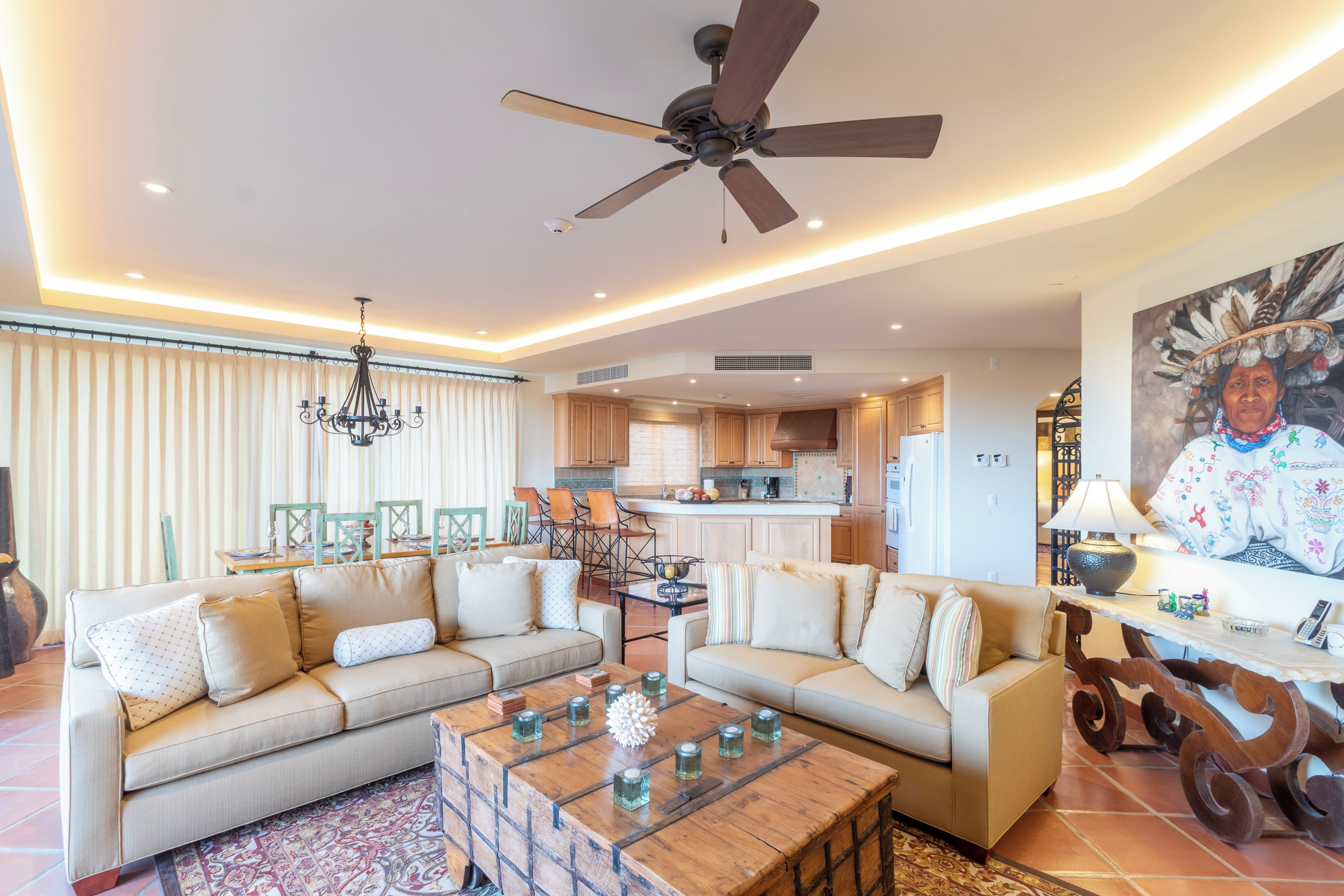 A spacious living room with a ceiling fan, a dining area with chairs, a kitchen with wooden cabinets, and a large painting on the wall.