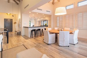 A spacious dining area with a wooden table and white chairs, a kitchen in the background, and a staircase leading to another level.
