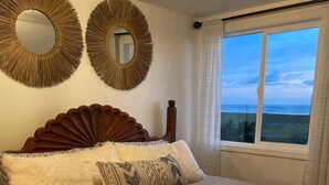 Ein Schlafzimmer mit einem großen Bett, zwei Wandspiegeln, einem Holz-Kopfteil und Blick auf das Meer durch ein Fenster.
