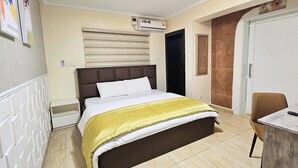 A hotel room with a bed, a desk, a chair, an air conditioning unit, and a door. - The Leneade - Mobo Apt. 6 at City Centre/Wuse 2 (Abuja)