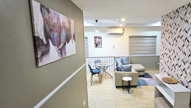 A modern living room with a sofa, a small round table, and a chair. There is a large painting on the wall and a patterned wallpaper on the right side. - The Leneade - Mobo Apt. 6 at City Centre/Wuse 2 (Abuja)