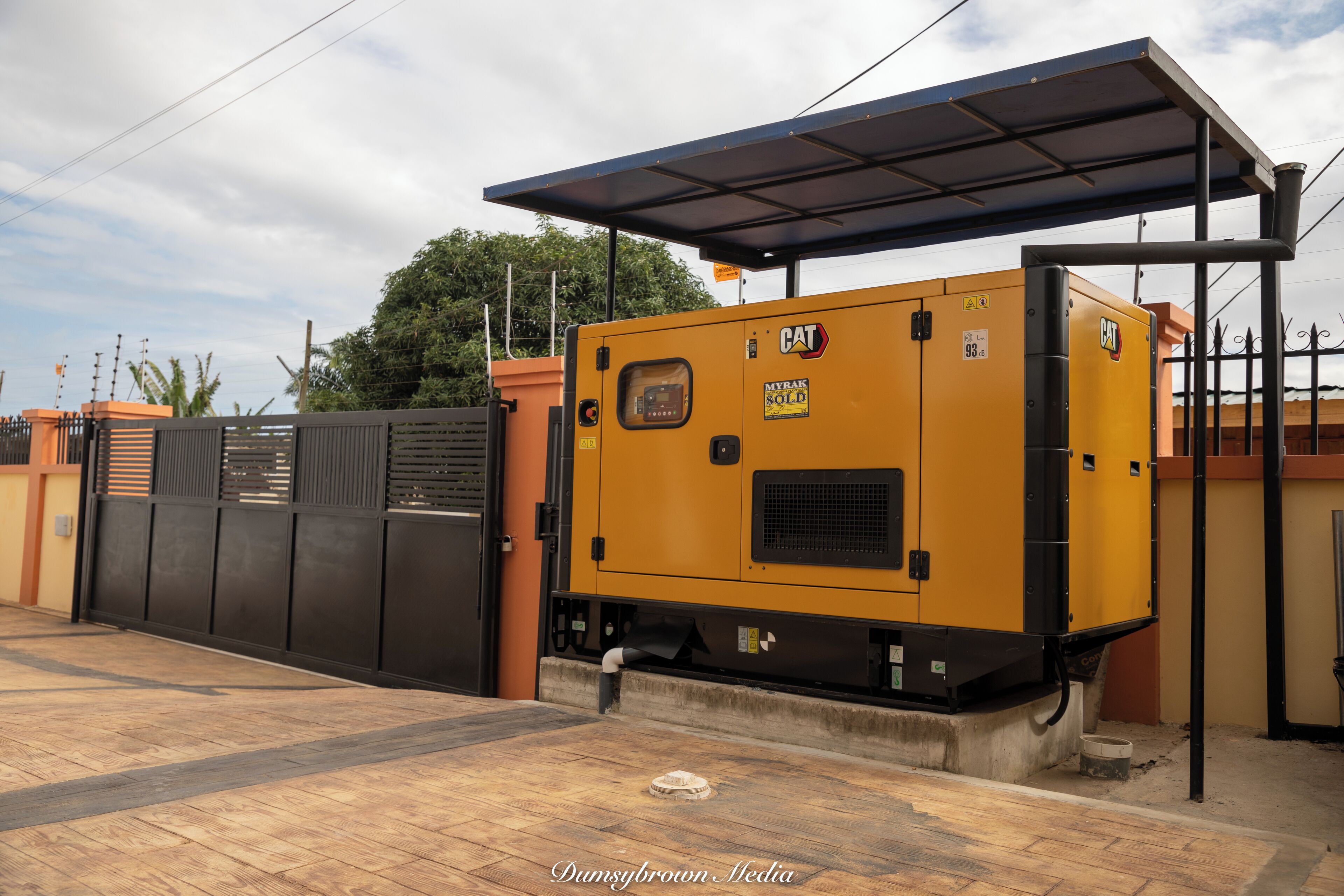 A yellow industrial generator with a roof structure, situated in an outdoor area with a paved floor and a metal fence in the background.