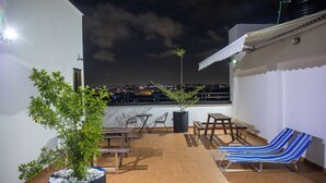 A rooftop terrace with a view of the city at night, featuring a potted plant, wooden furniture, and striped lounge chairs. - Deluxe 1 Bed Apartment - Wifi, Airon, Parking (Accra)