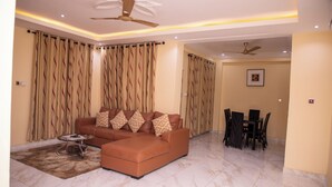 A living room with a brown sofa, a glass coffee table, and a dining area with a table and chairs. - Deluxe 1 Bed Apartment - Wifi, Airon, Parking (Accra)