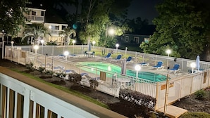 Vue nocturne d’un espace piscine avec une terrasse délimitée par une clôture, des chaises longues et un bâtiment en arrière-plan.