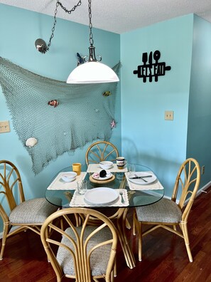 A dining area with a glass table, wicker chairs, and a blue wall with a net decoration. - Feel @ Home in Casita Cruz, Pool, 2miles beach, Laundry, King Bed & Free Parking (Myrtle Beach)