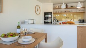 Una cocina moderna con una mesa de comedor de madera, sillas blancas y un cuenco de fruta. Hay un reloj en la pared y una botella de vino en la encimera.