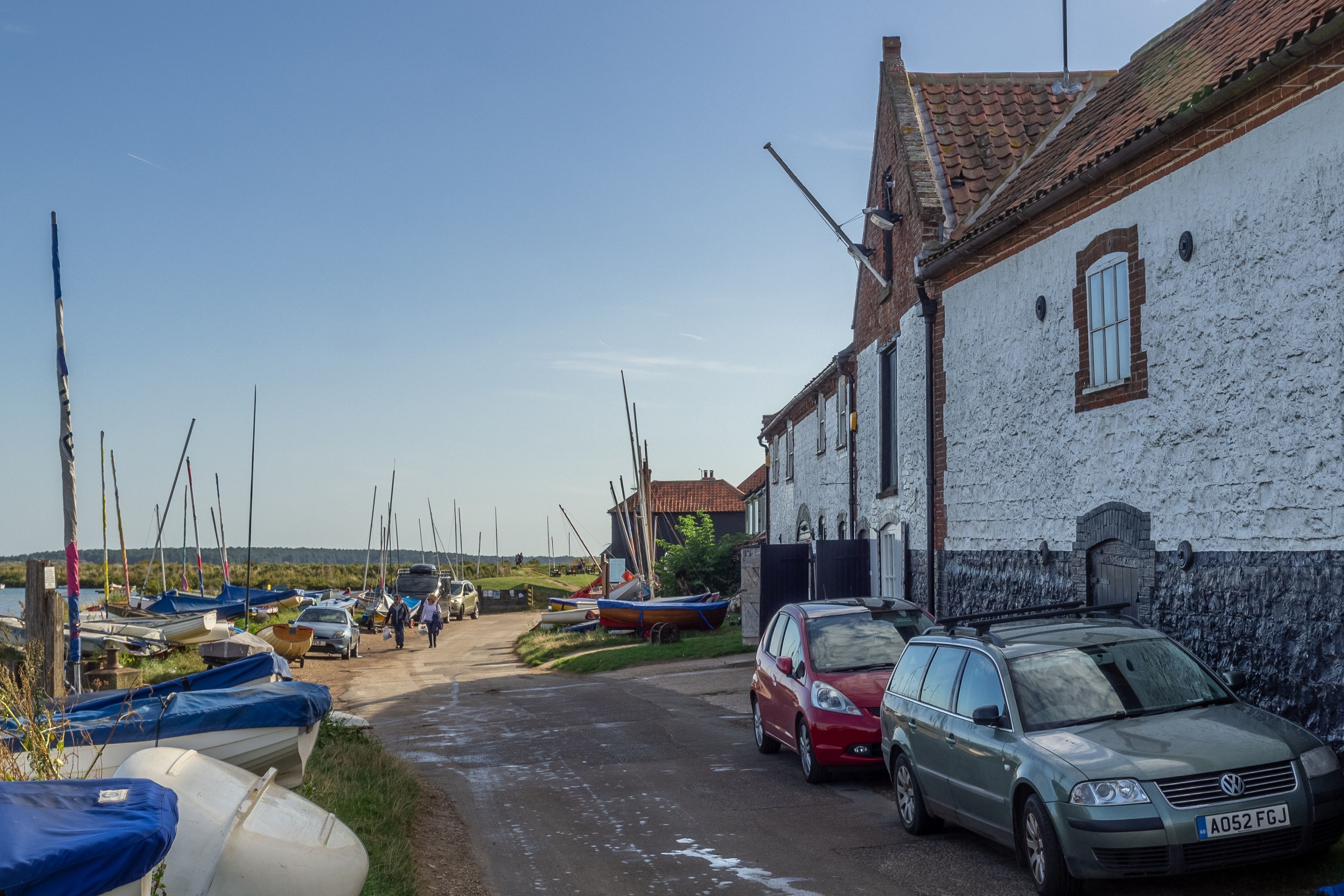 A narrow street with parked cars and boats, leading to a building with a white facade and a red-tiled roof.