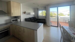 A modern kitchen with a microwave, stove, and wooden cabinets. There is a dining area with a table and chairs, and a balcony with a view of the city.