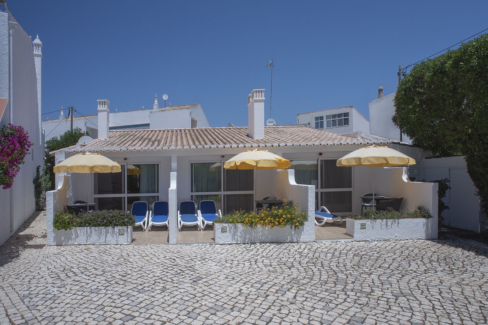 A courtyard with outdoor seating, including blue chairs and yellow umbrellas, surrounded by white walls and cobblestone flooring.
