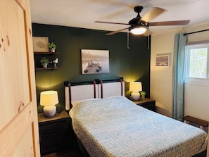 Een slaapkamer met een bed, twee nachtkastjes met lampen, een plafondventilator, een wandplank met decoratieve objecten, een ingelijst schilderij en een raam met gordijnen.