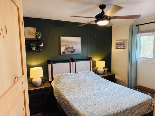 A bedroom with a bed, two bedside tables with lamps, a ceiling fan, a wall-mounted shelf with decorative items, a framed picture, and a window with curtains.