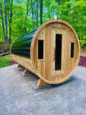 Een houten sauna met twee ramen en een gebogen ontwerp, gelegen op een grindpad met een bos op de achtergrond.