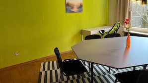 Una stanza con una parete verde brillante, un quadro, un tavolo con sedie, una finestra con tende e lampade a sospensione.