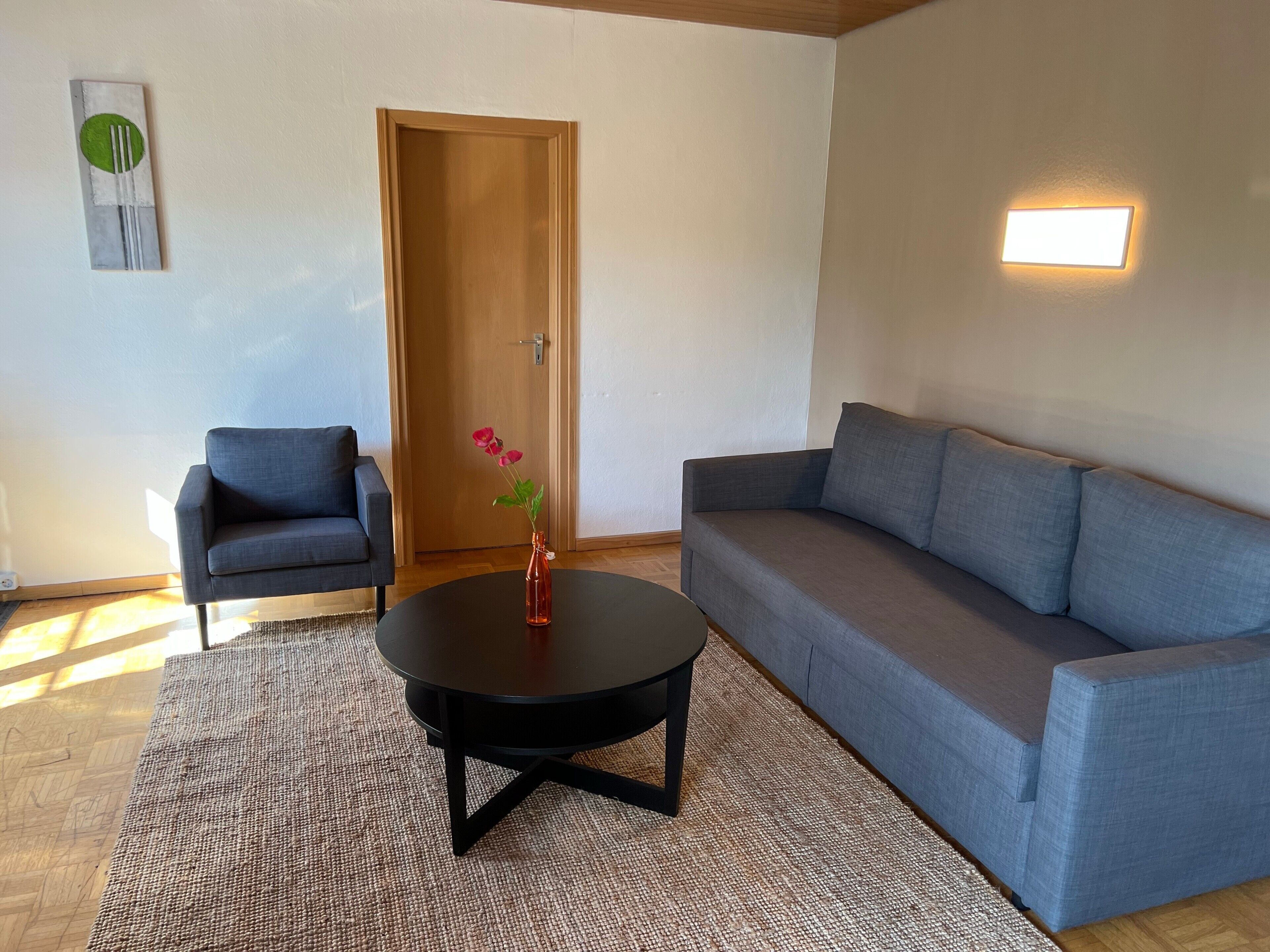 Uma sala de estar com um sofá cinza, uma mesa redonda pequena, uma poltrona azul, uma porta de madeira e um luminário fixado na parede.