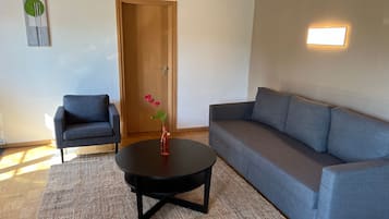 Uma sala de estar com um sofá cinza, uma mesa redonda pequena, uma poltrona azul, uma porta de madeira e um luminário fixado na parede.