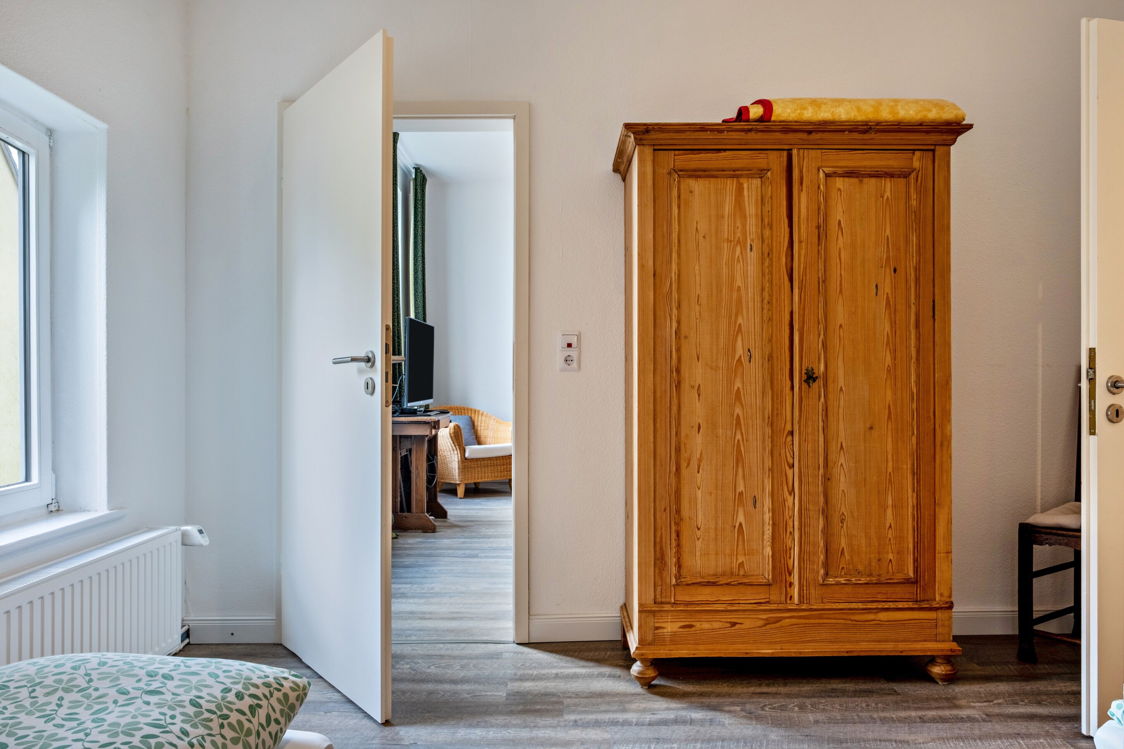 A room with a wooden wardrobe, a radiator, a window, and a door leading to another room.