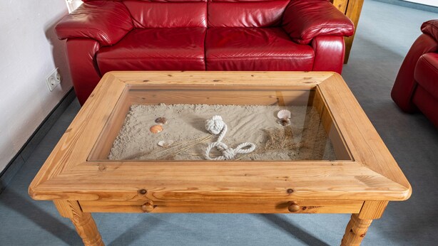 Une table basse en bois avec un plateau en verre et un matériau qui ressemble à du sable, un canapé en cuir rouge et un sol recouvert d’une moquette bleue.