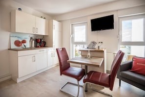 A modern kitchen with white cabinets, a dining table with two chairs, a flat-screen TV, and a sofa with a red pillow.