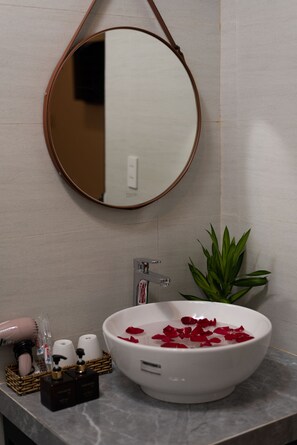 A bathroom with a round mirror, a sink with rose petals, a potted plant, and toiletries in a basket. - Tomorrow Villa Hoi An (Da Nang)