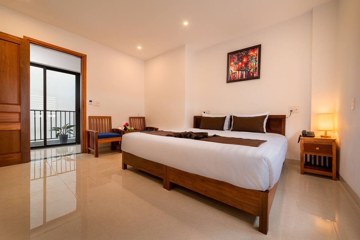 Ein modernes Schlafzimmer mit einem großen Bett, einem hölzernen Nachttisch, einem Sessel, einem Schreibtisch, einer Zimmerpflanze und einem Balkon mit Blick.