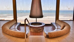 A room with a stone fire pit, a curved sofa, and large windows overlooking the beach. - Steps to Sand, Luxury Family Townhome with Central A/C BBQ Crib Resort Amenities (Solana Beach)
